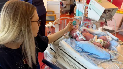 Family picture A baby lies in an intensive care cot, attached to multiple tubes and wires as his mother leans in to stroke his head.
