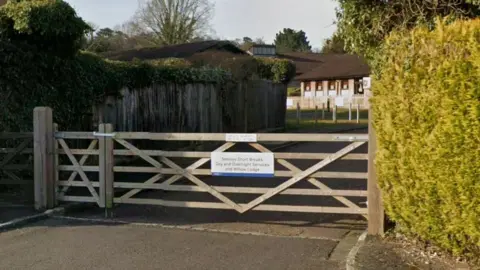 Google A wooden gate across a road, with a sign saying "Seeleys Short Breaks day and overnight services" in black lettering. There is a hedge to the right and a wooden fence to the left. A brick-built building with pitched roof is seen in the background.