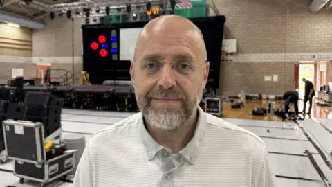 A bald man with facial hair and glasses. He is looking at the camera and standing in a sports hall covered in musical equipment.
