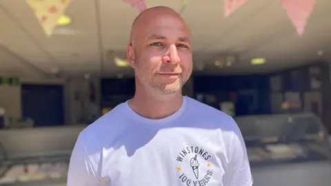 Tom Veal is wearing a white T-shirt with the words "Winstones, 100 years" on it, and smiles at the camera. The ice cream counter can be seen behind him.