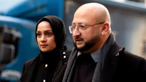 PA Media Soniya and Zaheer Ahmed are dressed in dark, formal clothing. Soniya on the left is wearing a black coat with decorative buttons and a head covering, while Zaheer is wearing a dark overcoat layered over a sweater and shirt, complemented by a scarf. In the background, there is a large blue vehicle.