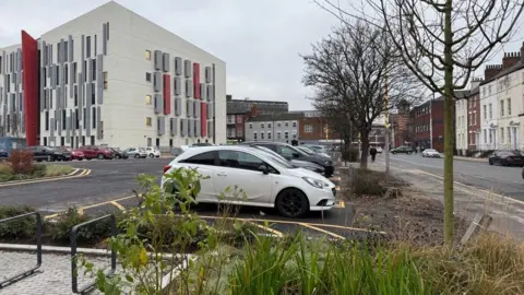 Cars parked in a car park next to a street with Georgian-style town houses to the right. A large modern building with grey and red panelling can be seen to the right.