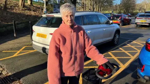 Nathan Turvey/BBC A picture of Kay Dobson wearing a peach coloured jacket standing next to her electric wheelchair with a car park behind her.