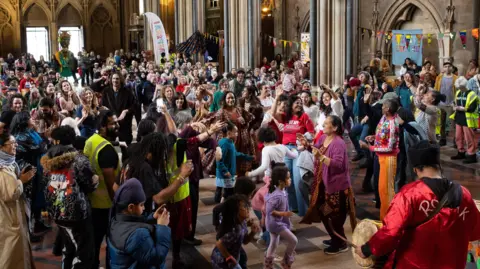 A large group of people dancing inside a church hall to a band. There is bunting across some of the internal archways in the church and there are lots of children among the dancers