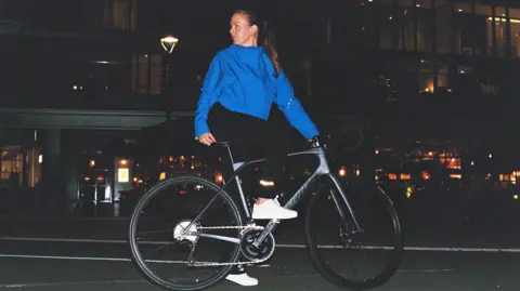A woman wearing black leggings with a reflective cuff, white trainers and a blue coat sitting on a bike with one foot on the ground, looking over her shoulder behind her. She's on a road and it's night, with a street lamp behind her.