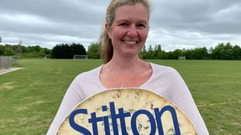 Sara Dunleavy is smiling at the camera standing on some grass and holding a sign saying "Stilton Cheese Rolling Festival" (only part of which can be seen). She has long, fair hair that is tied back and is wearing a striped top.