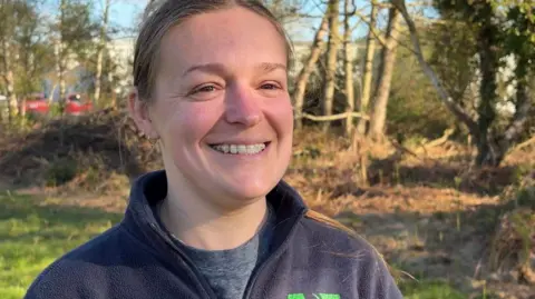 The picture shows a woman standing outdoors in bright, low sunshine that gives the scene a warm, early‑morning or late‑afternoon feel. She is smiling at the camera, wearing a dark fleece with a green logo on it, and her hair is tied back.