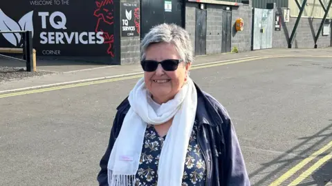 Sean Dunderdale/BBC A woman standing outside a football stadium and smiling at the camera. She has short grey hair and is wearing a floral blouse, an undone blue jacket, a white scarf and black sunglasses.