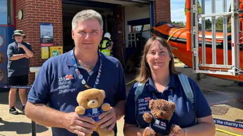 Allan and Helen Thornhill Two people wearing blue RNLI polo tops stood outside a lifeboat station, with an orange RIB lifeboat behind them. A man is stood behind them to the left