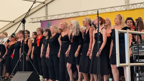 Art Asia A large group of women wearing black dresses are stood together on a stage during a performance.
