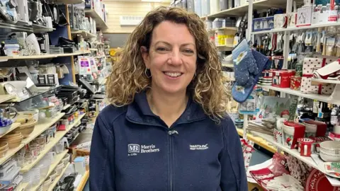 A smiling Katherine John standing in her shop looking directly at the camera wearing a navy blue zip-up top with MB Morris Brothers printed on it in white lettering . Either side of her are shelves packed with various products for sale, including oven gloves, spoons, glass measuring jugs and see-through plastic containers.