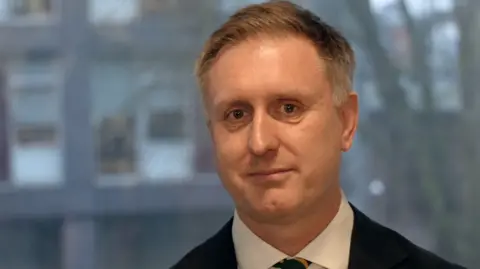 Vikki Irwin/BBC Ed Garratt standing inside a building in front of a window. He is wearing a suit with a black suit jacket and tie. He is looking directly at the camera and smiling.