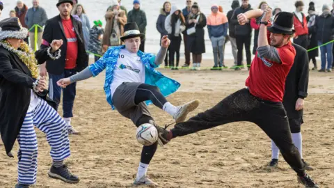 Men in fancy dress playing football on Scarborough beach with spectators watching.