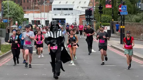 A person dressed as Darth Vader from Star Wars is running in front of other men and women, who are also competing but wearing shorts and T-shirts.