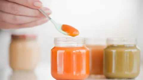 Getty Images hand holding spoon with Orange coloured baby food, beside jars of Orange and brown baby food