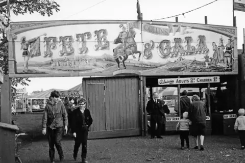 Walsall Archive A black and white image of ann admission booth for Pat Collins Fete and Gala. People are queuing to pay their money beneath a fairground sign. Two boys can be seen walking in the opposite direction. 