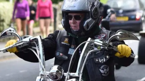 Paul Perry A man riding a motorcycle with shoulder-height handle bars. He is wearing a helmet and a jacket with badges on it.