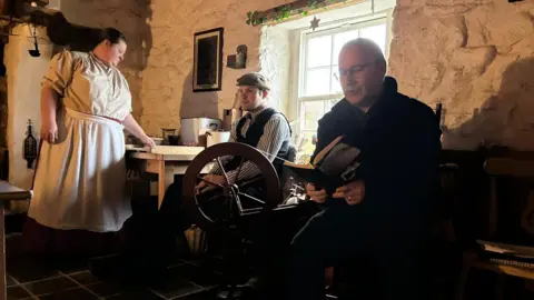 On the right, a man with shorn grey hair and glasses reads poetry from a book, sitting down in black clothes.
To his right is an old spinning wheel for yarn or thread, and a young man seated dressed in a flat cap and striped white shirt under a black waistcoat.
A woman with black hair tied back neatly stands to the left dressed in an old fashioned white blouse and apron, making soda bread from scratch.
Everyone is inside the cottage, as the window draws a white light in behind the young man.