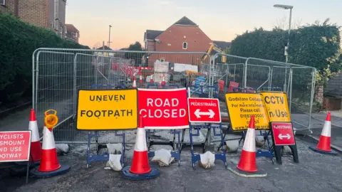 Mark Carter/BBC Wire fences surround a roadworks side. Multiple red and yellow warning signs and some traffic cones have been set up in front of the fences.