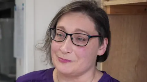 Tom Ingall/BBC A woman with cropped brown hair wearing rectangular glasses looks away from the camera