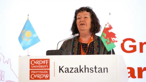 Cardiff University A woman with shoulder-length brown, wavy hair at a lectern which says Cardiff University Kazakhstan and has the Welsh and Kazakhstan flags on it.