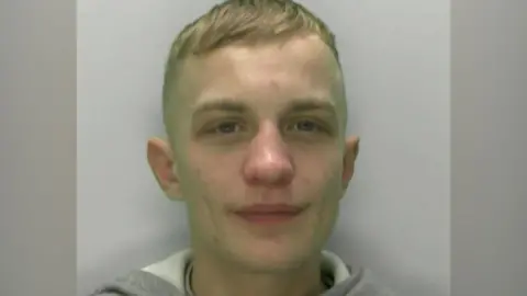 Jack Bradley appears to be smiling slightly in his police mugshot as he stares just off-camera. He is standing against a grey-white background, wearing a grey hoodie with a white inside. He has short blond hair.