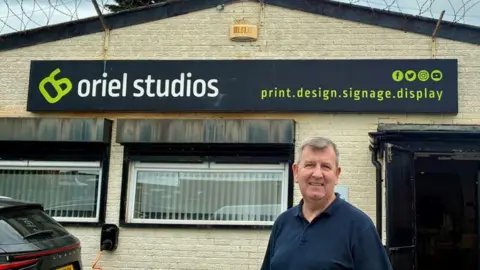 LDRS Man stands in a navy-blue polo shirt in front of a building which has a sign reading 'Oriel Studios'.