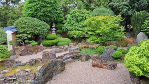 Japanese Garden Society A view of a Japanese style garden featuring a low stone wall, stone lantern and bridge balanced on rocks over gravel representing water. Small ornamental trees are set around rocks.
