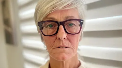 Jackie McTaggart is looking directly at the camera. She is wearing a cream knitted jumper and has large brown rimmed glasses. She has short white hair. 