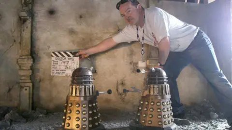 Mike Tucker Mike Tucker holds a clapper board that says "Doctor Who" close to two model Daleks, as they prepare to film an effects shot for Day Of The Doctor.