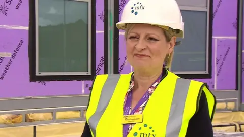 BBC Professor Karen Dunderdale. She is standing outside one of the centres. She is wearing a white hard hat and a fluorescent jacket