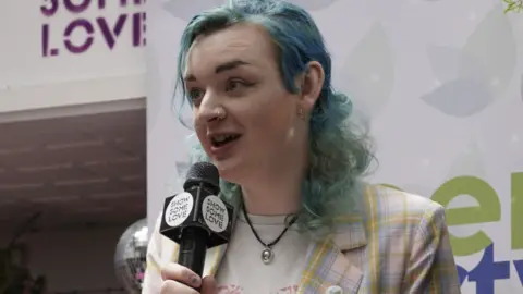 Green Party NI Ash Jones holding a microphone that says "show some love", wearing a pink, lilac and yellow tartan jacket