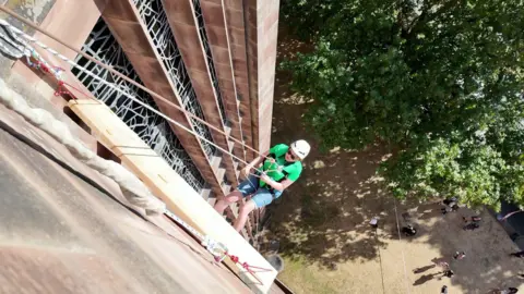 A person in light denim shorts and a green top wearing a white helmet is abseiling down the wall of a large brown brick cathedral