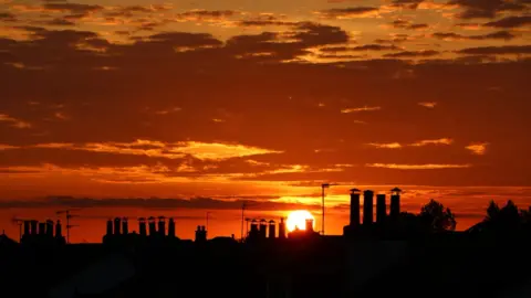 A golden sunset with silhouetted rooftops and chimneys in the foreground
