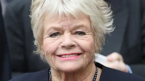 Judith Chalmers smiling in a navy blue blazer and necklace in 2016
