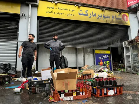 Pacemaker Two shopkeepers outside fire-damaged store on Donegall Road with damaged stock strewn across the pavement