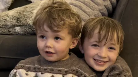 Ella Two young boys in brown patterned jumpers are hugging and smiling at the camera. They are sitting on the floor in front of a brown sofa
