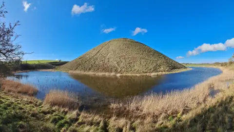 WeatherWatchers/Sonia Silbury Hill - which is an almost symetrical mound - standing above flood water and with a very blue sky in the background. The flooding looks like a moat, with the hill reflecting in it.