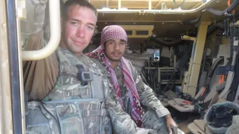 Gareth Evans A man in a camoflage waistcoat and army combats inside a tank, next to a man also in camo but wearing a red and white checked headscarf