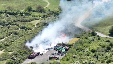 Eddie Mitchell Thick smoke pours from a concrete square where there are scores of wheelie bins. 