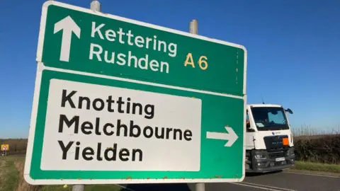 Alex Pope/BBC A large green and white road sign, with different place names on, with a lorry driving along the road. A blue sky can be seen above, a grass verges either side of the road. 