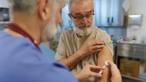 a hospital doctor with grey hair and a beard is wearing blue clothing and giving a vaccine jab to a man with grey hair and beard who is wearing glasses