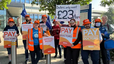 Resident doctors stand on a picket line outside Hull Royal Infirmary. They are holding orange and white signs featuring the BMA logo. The hospital building is visible in the background.