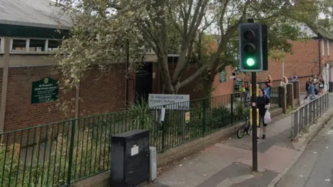 A street view of St Margaret's CEVA Primary School. It is a low-lying 1950s or 1960s looking brick building, with two signs with its name on it. There is a black metal fence between the school boundary and the pavement and people are clustered by its gate in the distance. 
