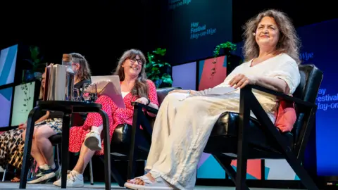 Adam Tatton-Reid/Hay Festival Jones sits alongside Beckerman on stage at Hay Festival. Jones, on the right, she wears white linen trousers and a white blouse. Beckerman wears a red and white polka dot jumpsuit and has long brown hair. She looks and smiles at Jones.