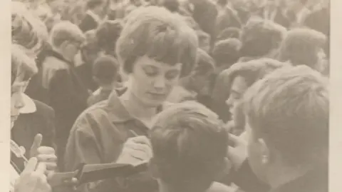 Anne Grimes Anne Grimes is pictured signing autographs in 1958 in Holland after a Corinthians game. She is surrounded by crowds of fans. 