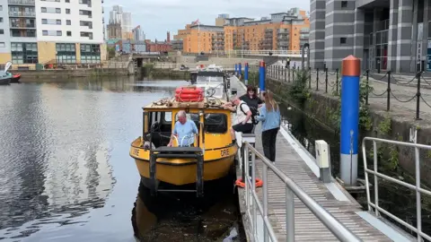 Dan Bater/BBC News People getting on a bright yellow river taxi in Leeds