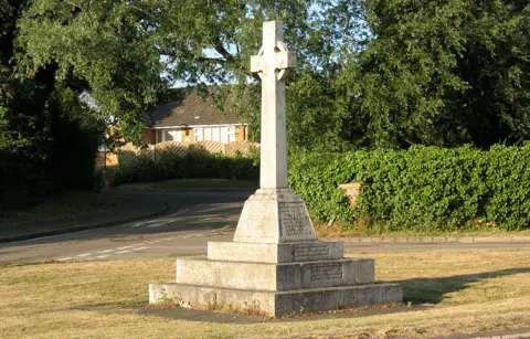 Stephen Craven/Geograph War memorial on a lawn, with road junction, hedging, trees and a bungalow in background 