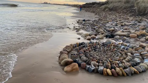 A coastal artwork made up of hundreds of coloured stones spiralling outwards is pictured on Cambois beach - you can see Lynmouth power station in the distance 