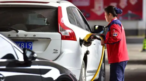 A female petrol station attendant refuels a white car
